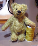The finished Norman teddy bear standing by a reel of thread to give a sense of scale. Norman is a sweet gold little teddy with black eyes and a black nose.