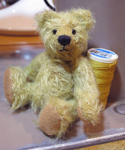 The finished Norman teddy bear sitting by a reel of thread to give a sense of scale. Norman is a sweet gold little teddy with black eyes and a black nose.