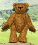 Treacle, a brown teddy bear made from the Treacle teddy bear kit standing on a wooden platform with a scenic background of trees and mountains.